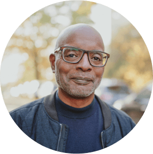 Man (actor portrayal) with glasses standing outside, headshot